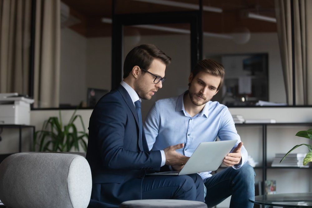 Two men in office setting looking at laptop, one in a suit, one in button-down, both focused.