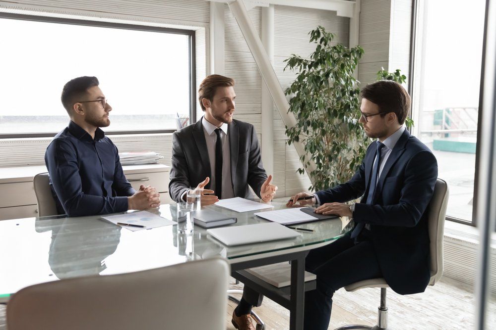 Three men in suits at a conference table discussing documents in a bright office.