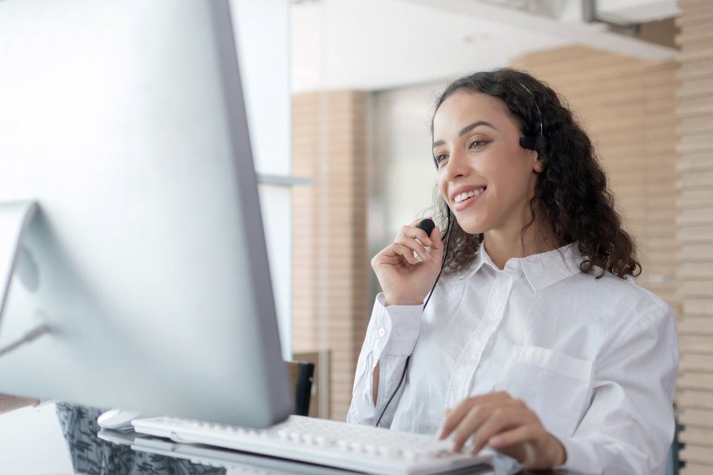 Woman with headset smiles while working on a computer, bright room.