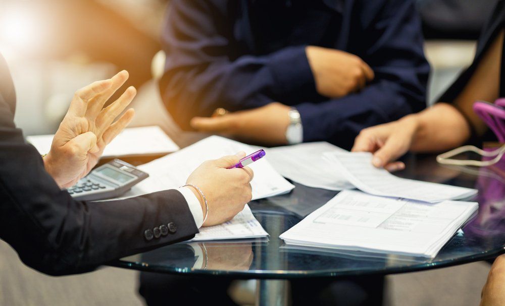 Business people at a table, discussing documents with gestures. Sunlight and a calculator are visible.