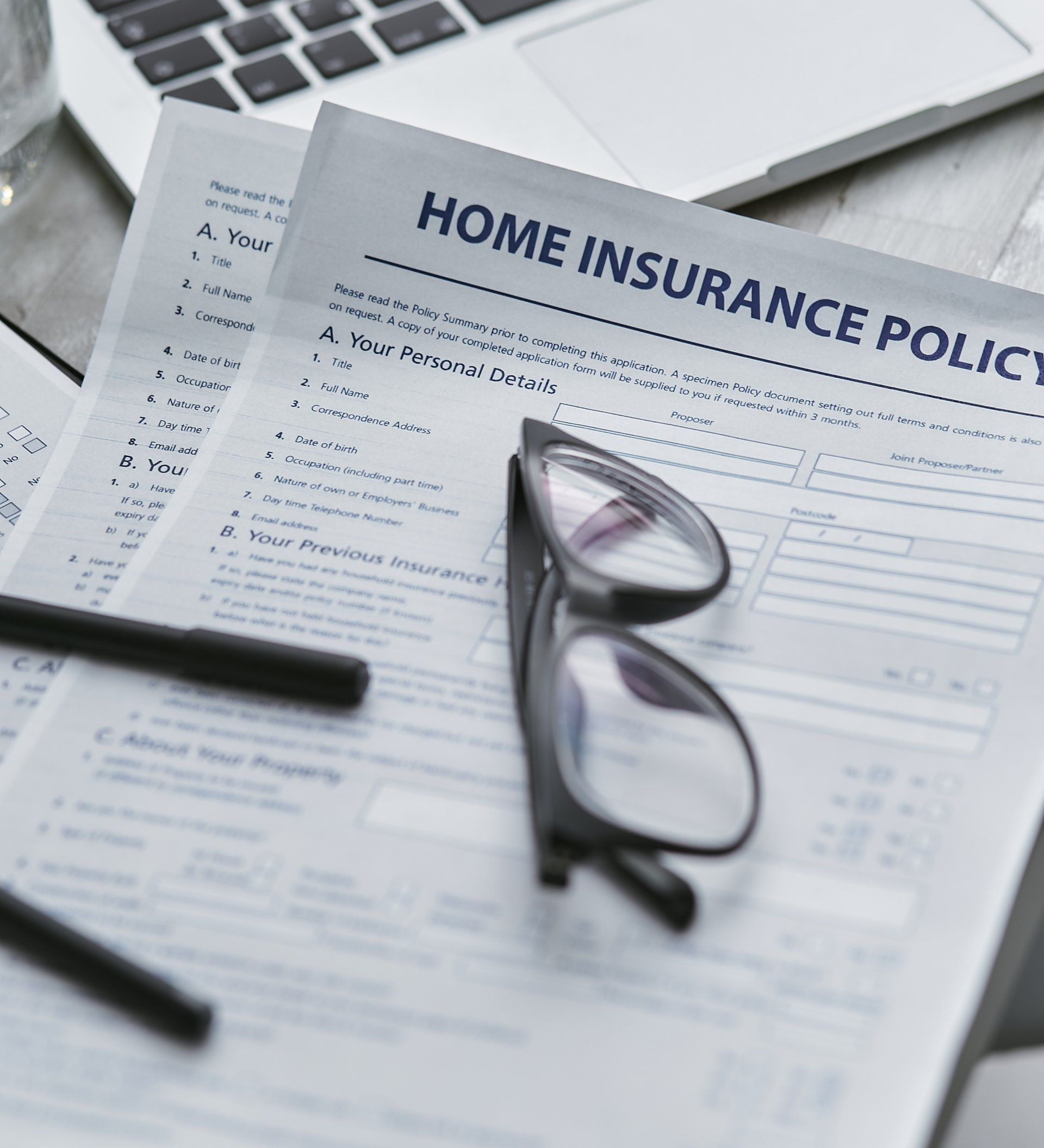 Home insurance policy document with eyeglasses and pen on top of a desk, near a laptop.