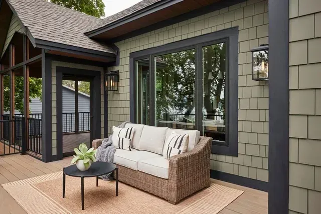 Outdoor patio with wicker sofa, coffee table, and greenery, set against a green shingled house.