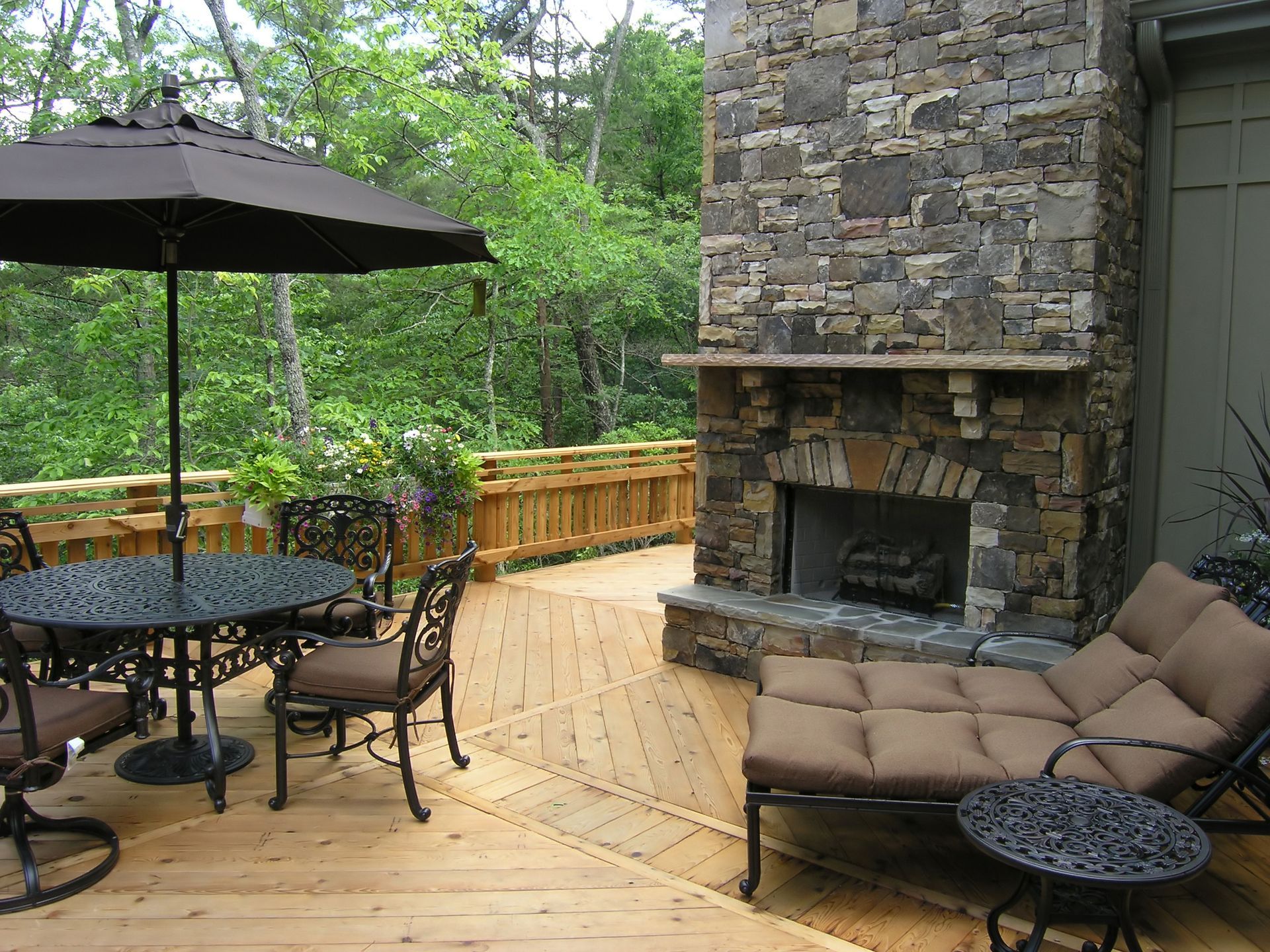 Patio with outdoor fireplace, seating, and umbrella, overlooking trees.