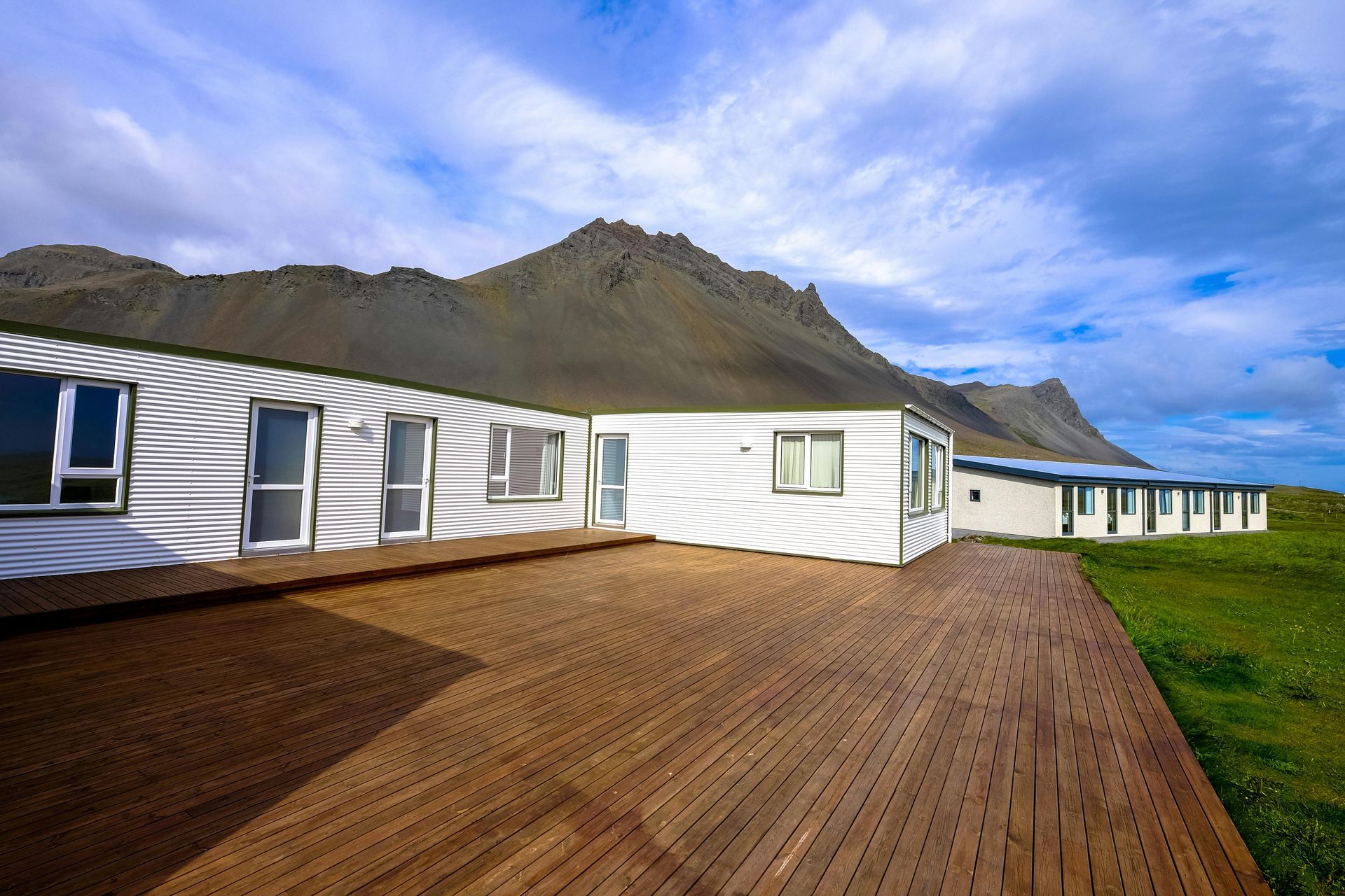 White buildings with wooden deck, mountain backdrop under cloudy blue sky.