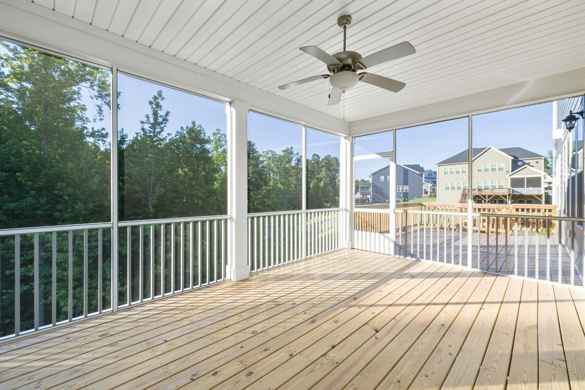 Screened porch with wooden floor, white railing and ceiling, open to view of trees and houses.