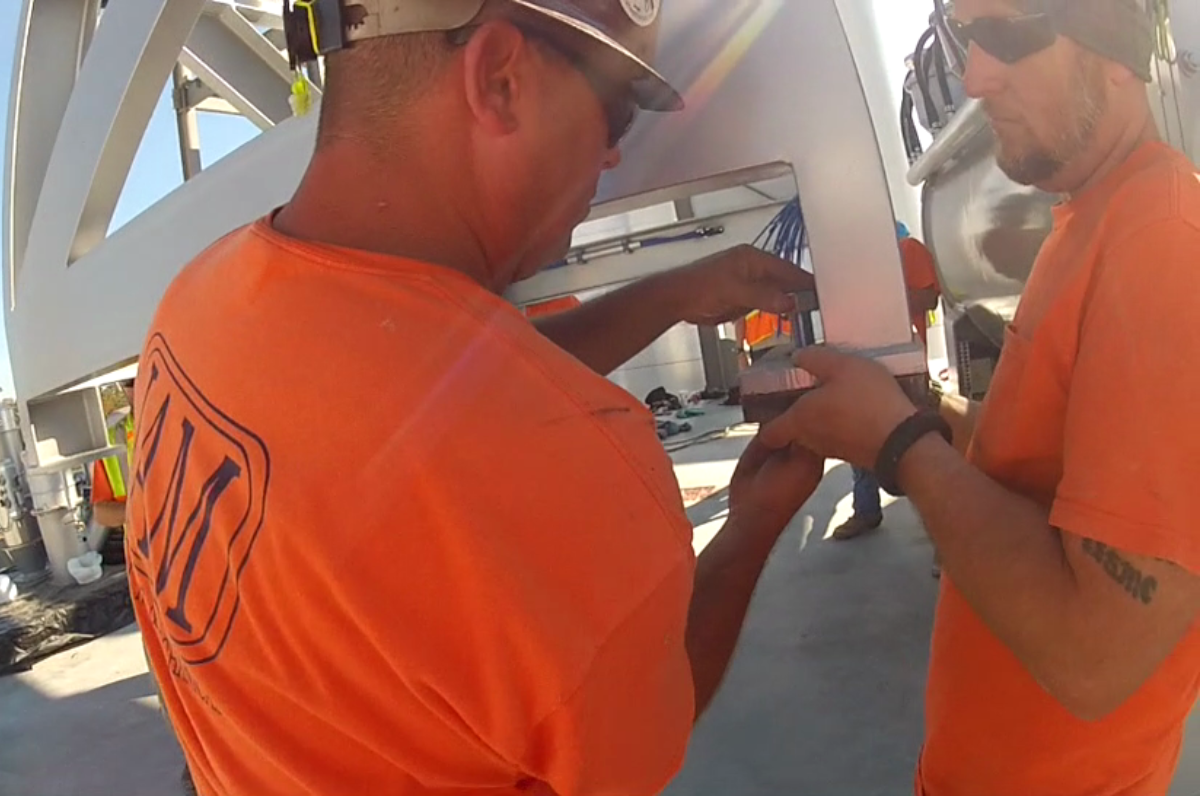 Two men in orange shirts are working on a metal structure.