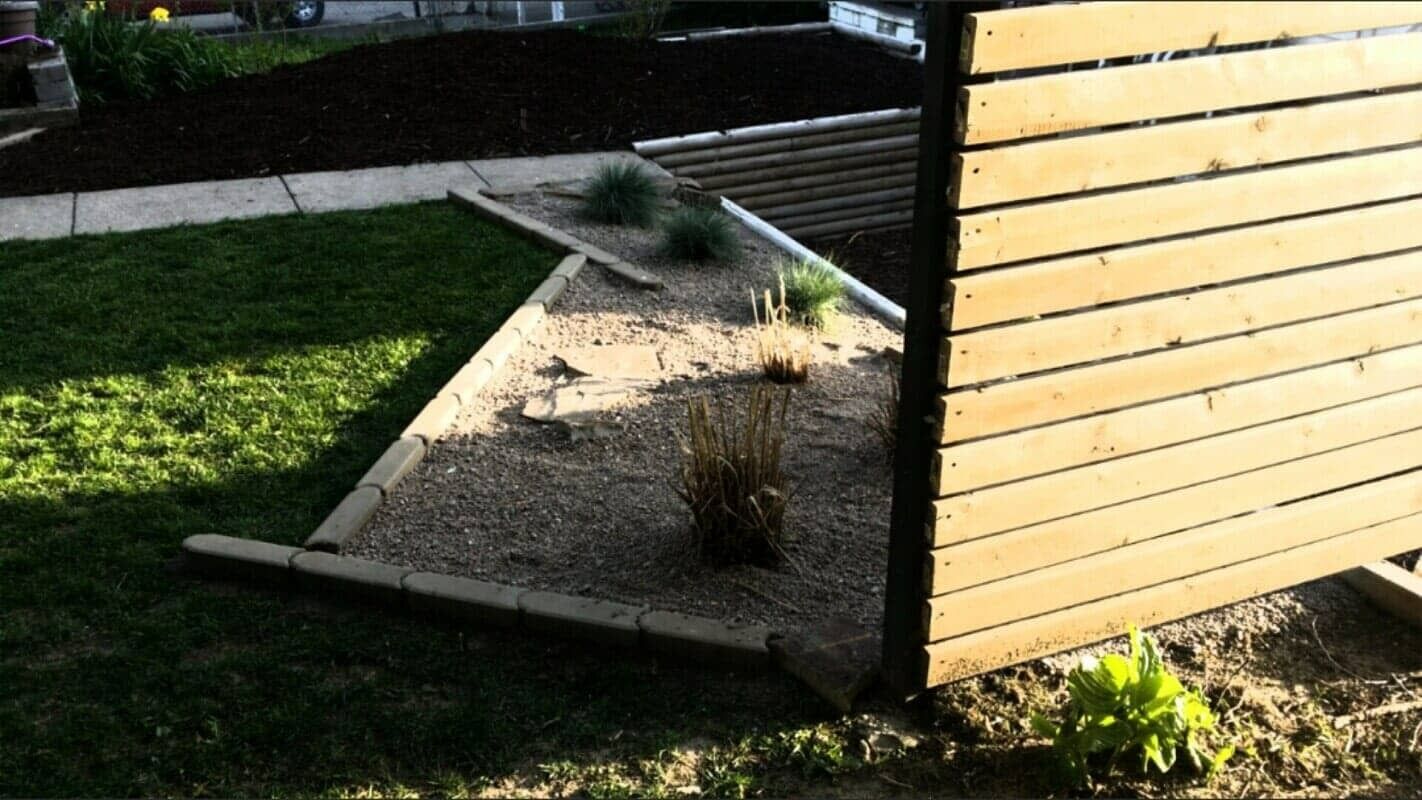 Raised gravel garden beds beside a yellow wooden building, with a green lawn in the background.