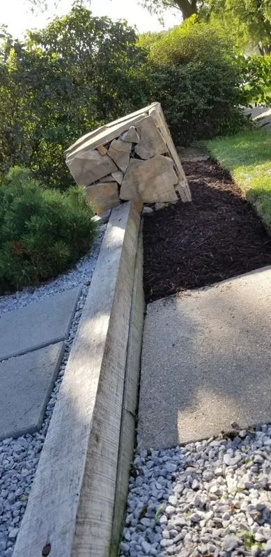 Damaged roadside retaining wall with a collapsed stone section beside a sidewalk and gravel landscaping.