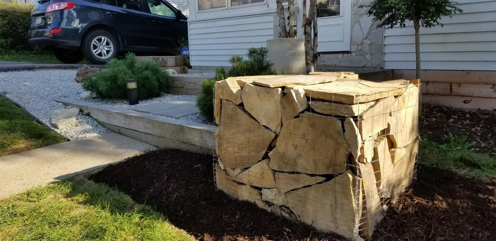 Large stone planter cube in a front yard beside a house, with a parked car in the background.