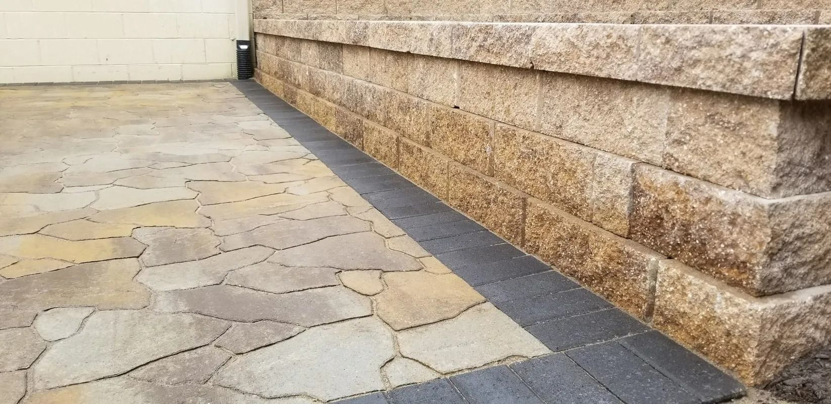 Stone walkway beside a tan textured wall, with cracked paving and a dark border strip