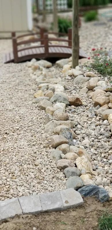 Rock garden with pale gravel, scattered stones, a small wooden bridge, and plants beside a path.