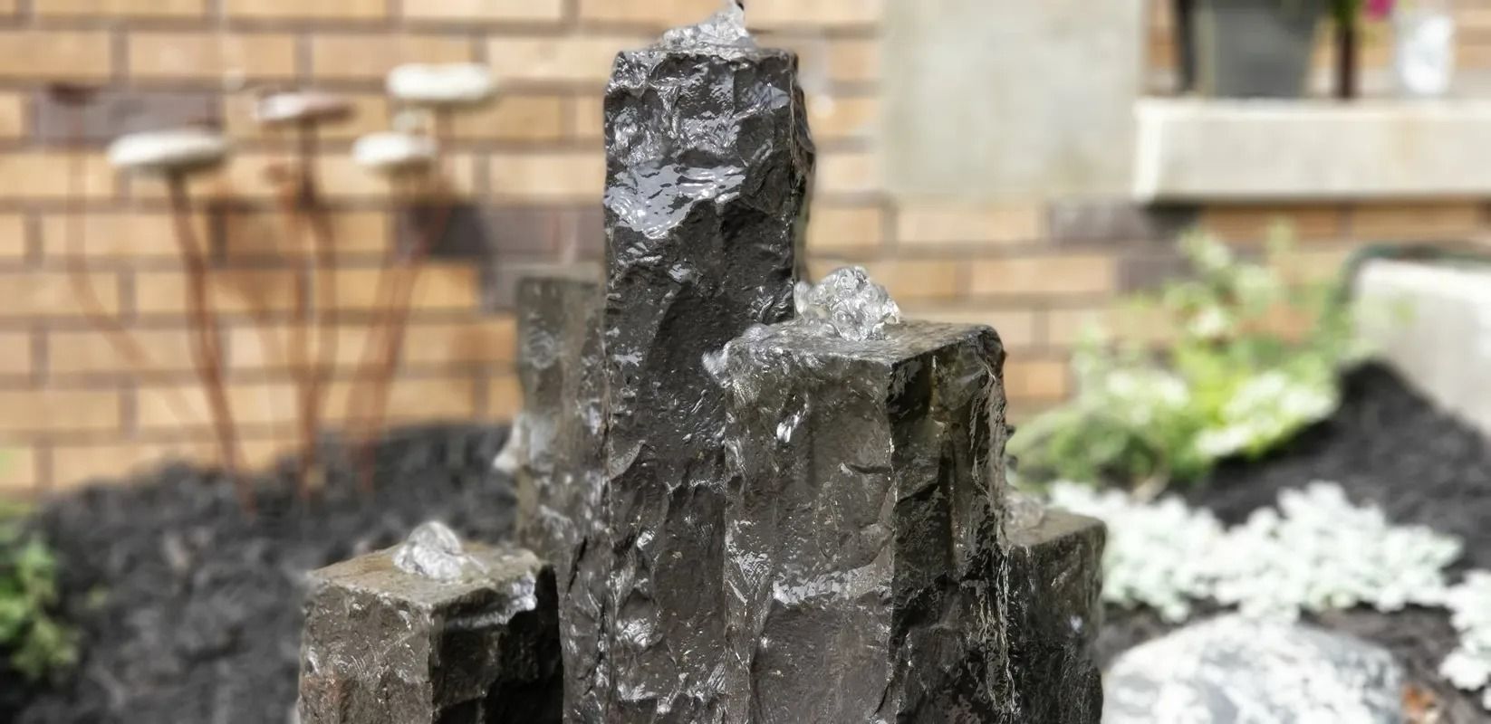 Dark stone fountain or sculpture with water dripping, set in a garden against a brick wall.