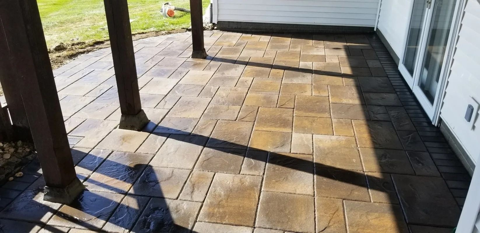 Sunlit stone patio beside a house, with dark posts and a grassy yard beyond