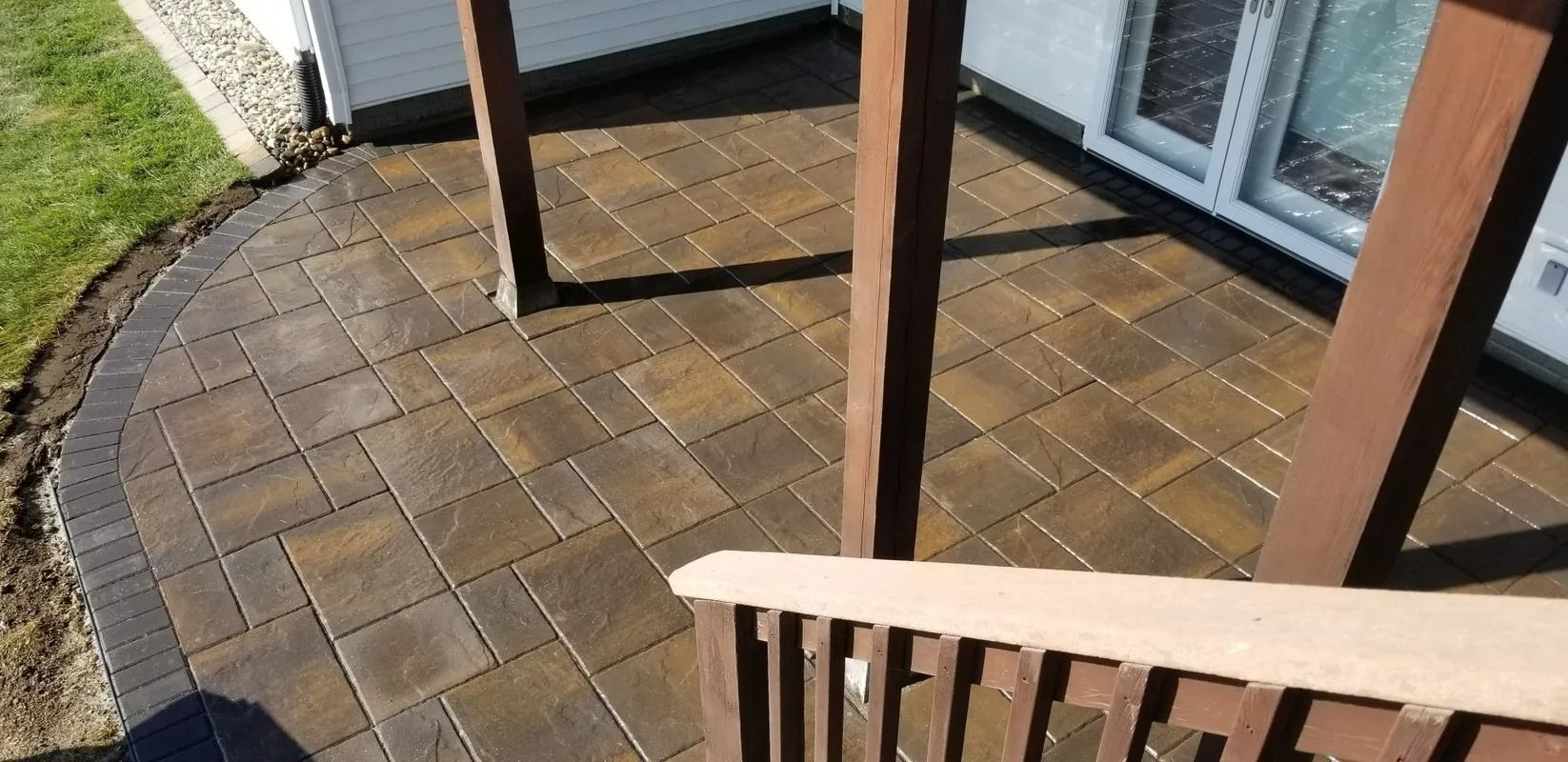 Patio with brown pavers beside a house, viewed from a wooden deck railing