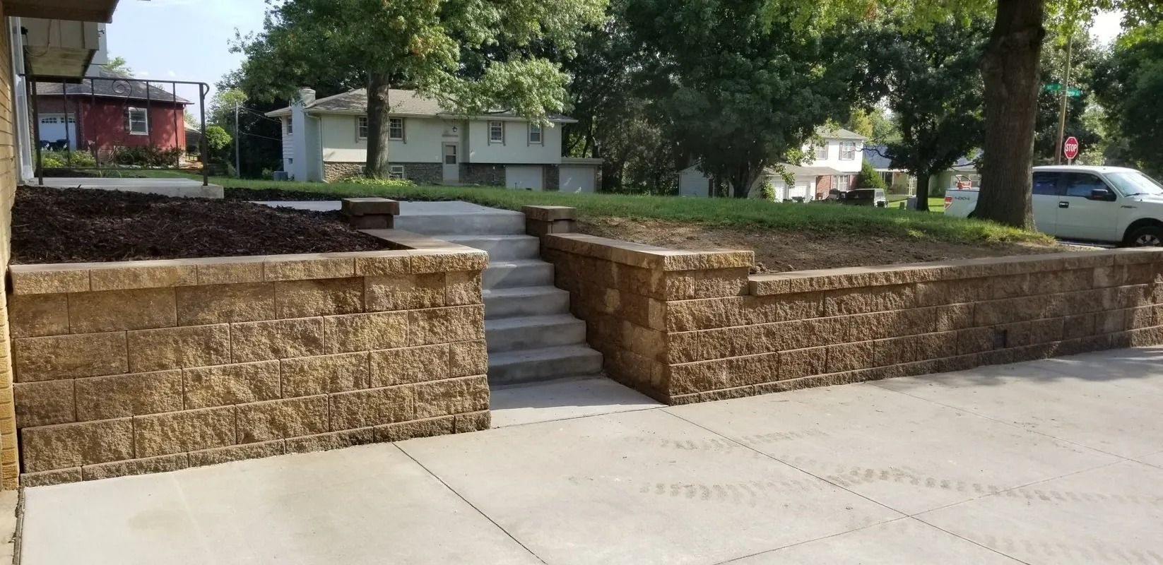 Stone retaining walls and steps leading to a raised residential walkway with trees and houses in the background