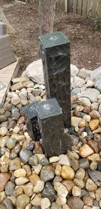 Two dark stone fountain posts among river rocks in a backyard garden, with water trickling down one side
