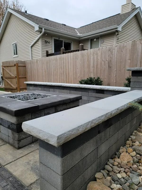 Backyard stone seating walls and fire pit in front of a house, with a wooden fence and rock landscaping