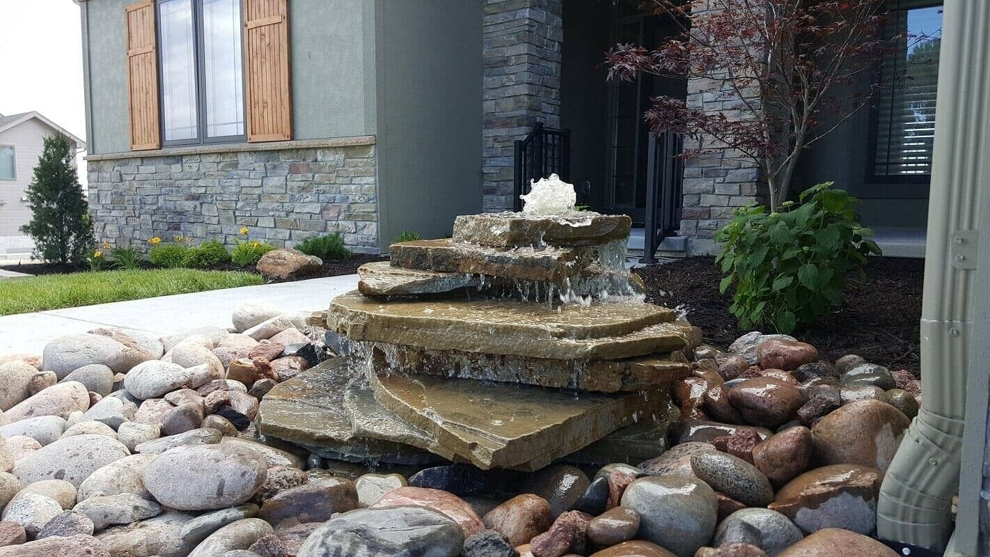 Stone fountain in a landscaped front yard with rocks and a house exterior behind it