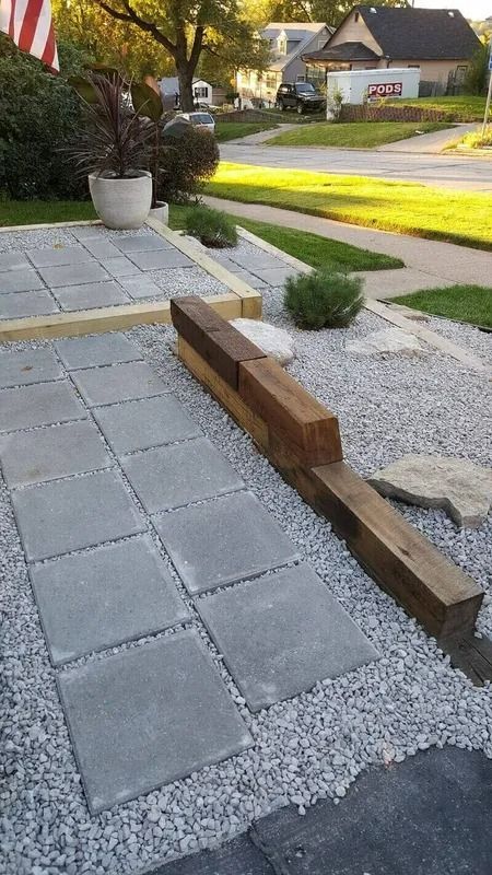 Front yard with gray pavers, white gravel, wooden edging, and a small shrub beside a lawn.