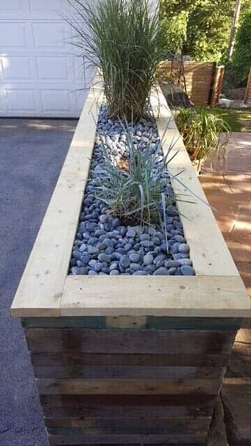 Tall rectangular planter with ornamental grasses and blue-gray stones beside a garage and patio.