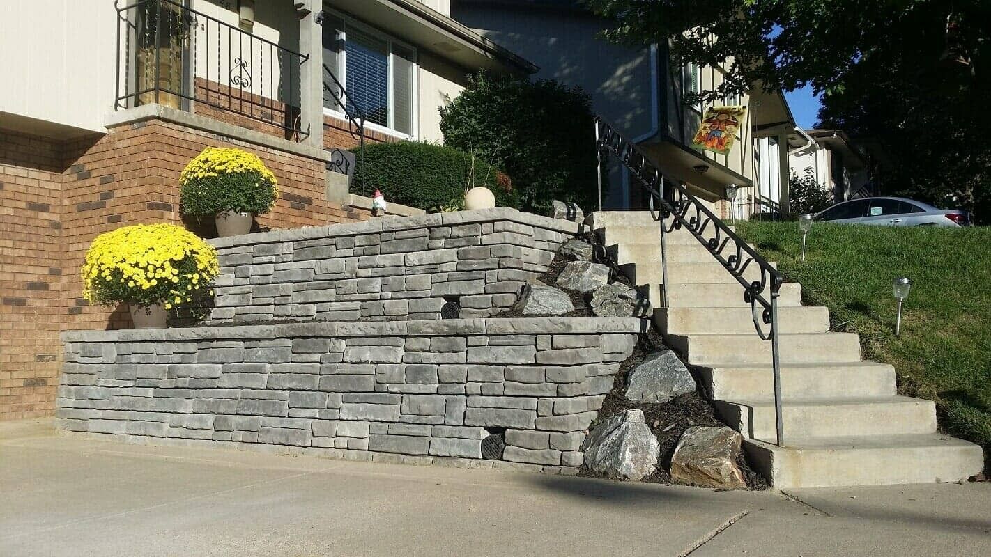 Stone retaining wall beside concrete stairs leading to a brick house, with yellow flowers and black railings.