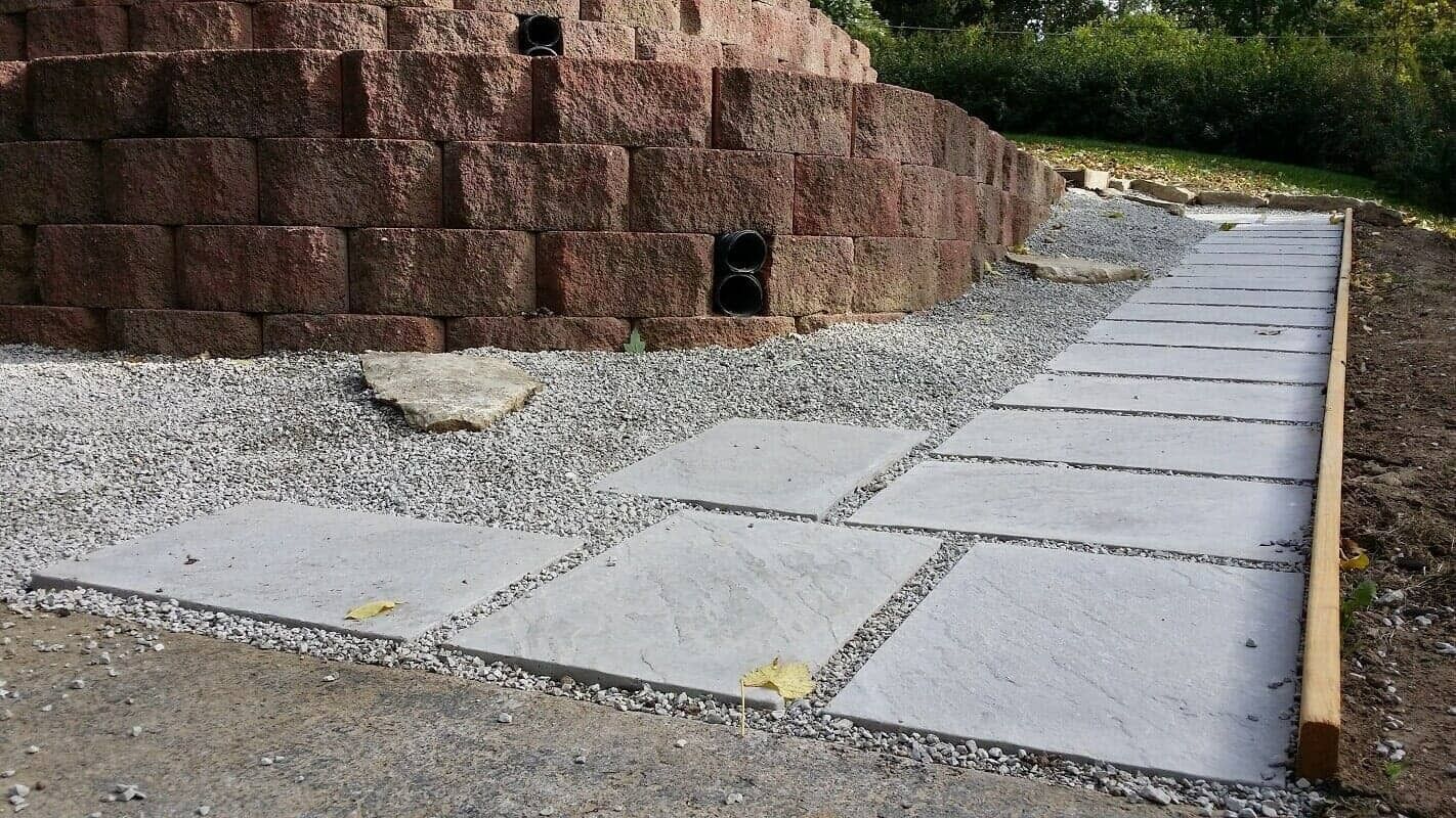 Concrete path beside a brown retaining wall, leading into a gravel area and greenery