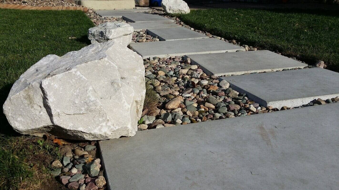 Concrete walkway with a large white boulder beside stepping stones and decorative gravel in a yard