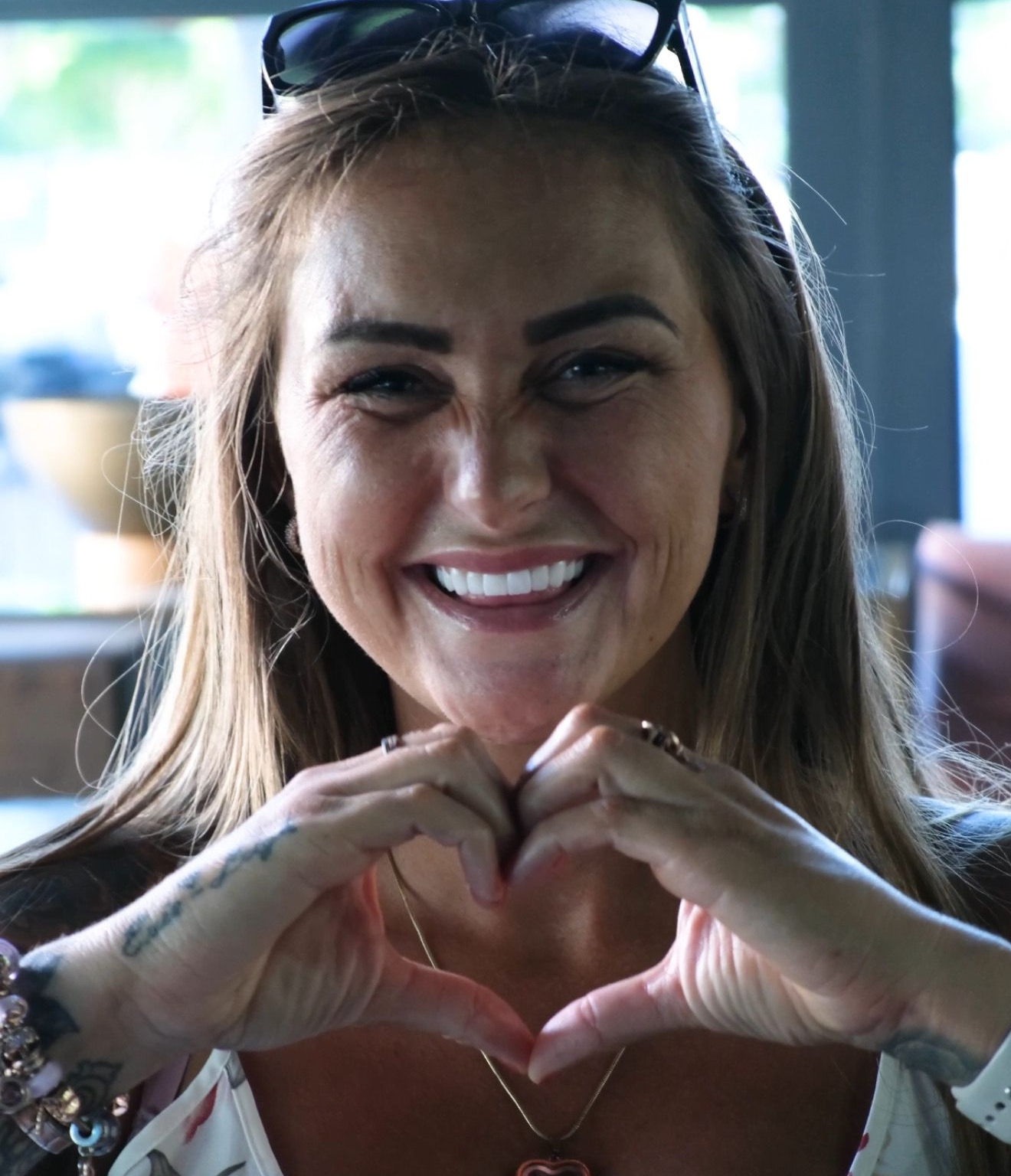 Woman smiles and forms a heart with her hands. She is wearing a floral top and necklace.