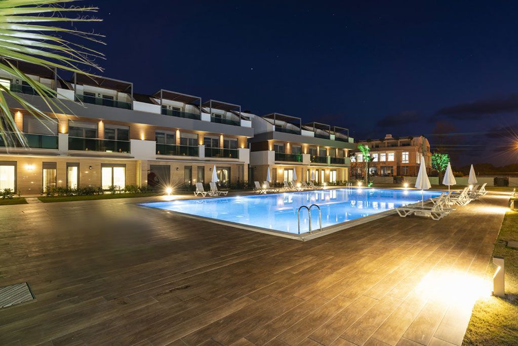 A pool at night with illuminated buildings and lights; palm tree on the left.