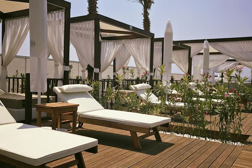 Beachside cabanas with white curtains and lounge chairs on a wooden deck, sunny day.
