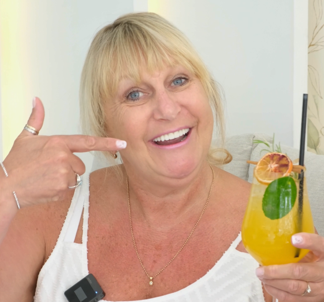 Woman pointing at an orange cocktail with a smile, wearing a white top.