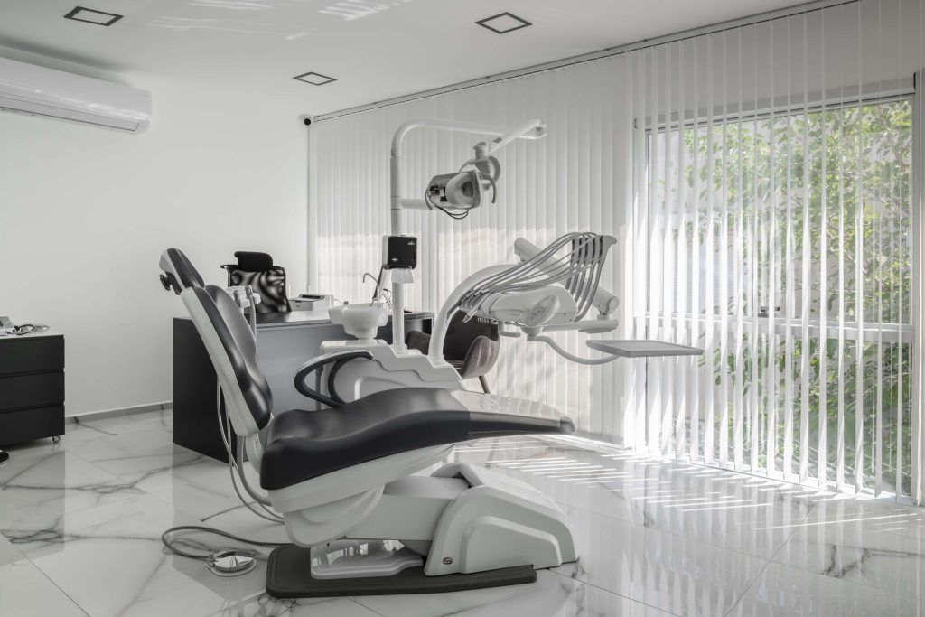 Dental office interior with chair, equipment, and window with blinds. White and gray color scheme.