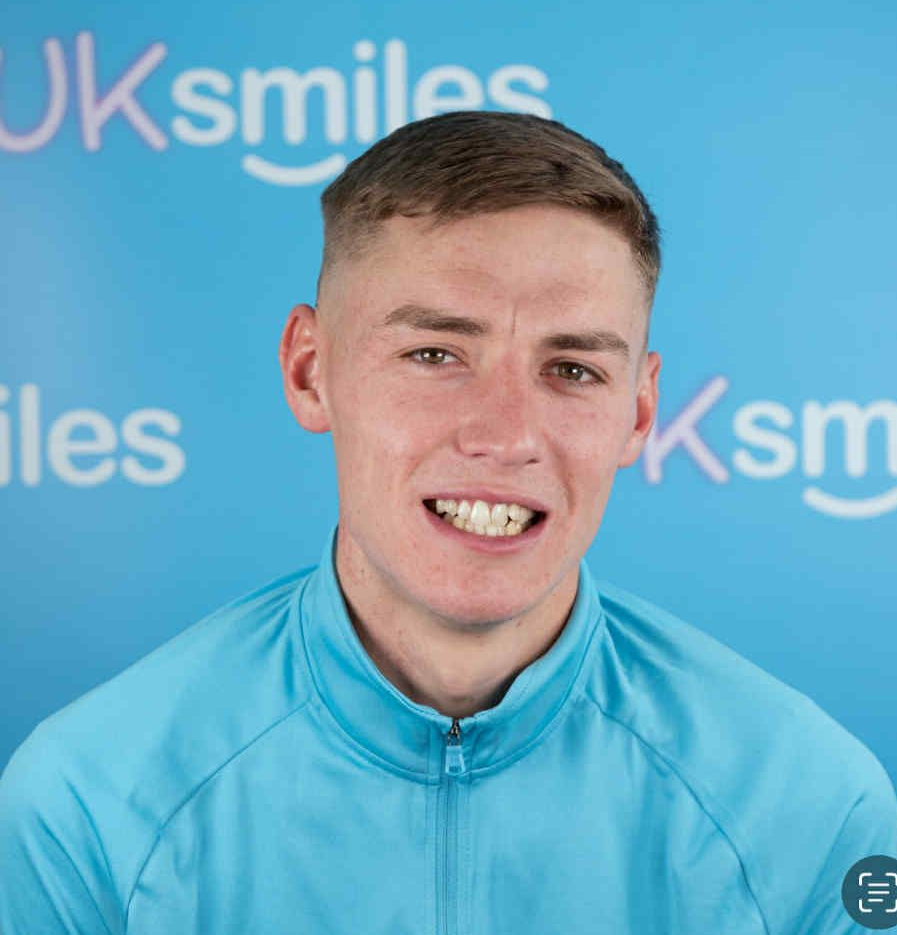 Man with short brown hair, wearing a blue athletic jacket, smiling in front of a blue background with text “UKsmiles.”