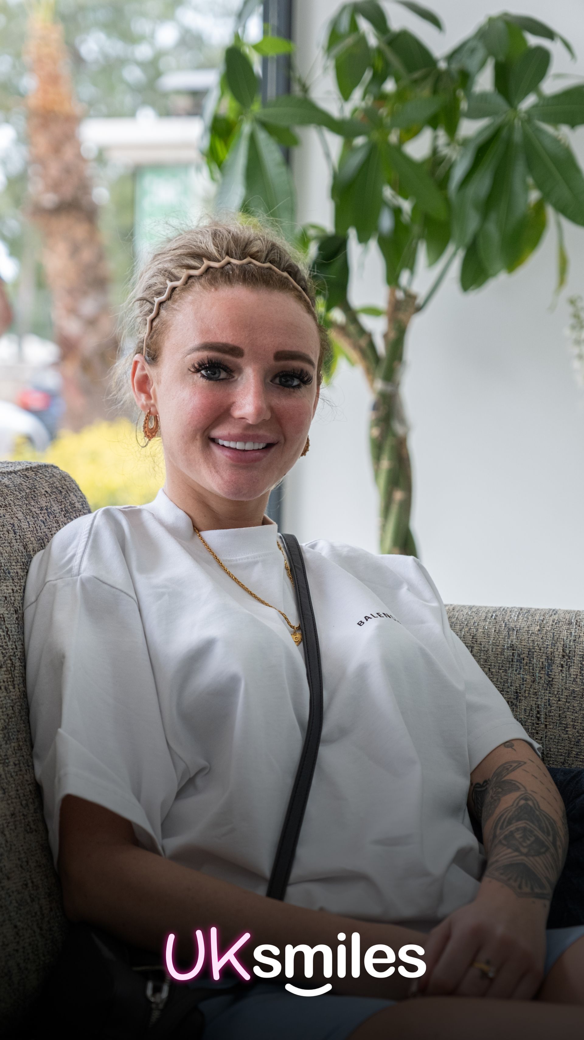 Woman smiles, seated indoors. Wearing white shirt, gold jewelry, and a headband. Tattoo on arm.