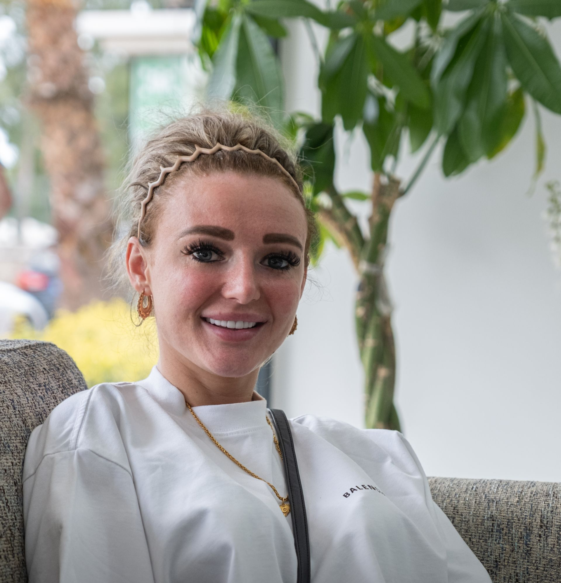 Woman with blonde hair smiles while sitting on a couch in front of a tree, wearing a white shirt and gold jewelry.