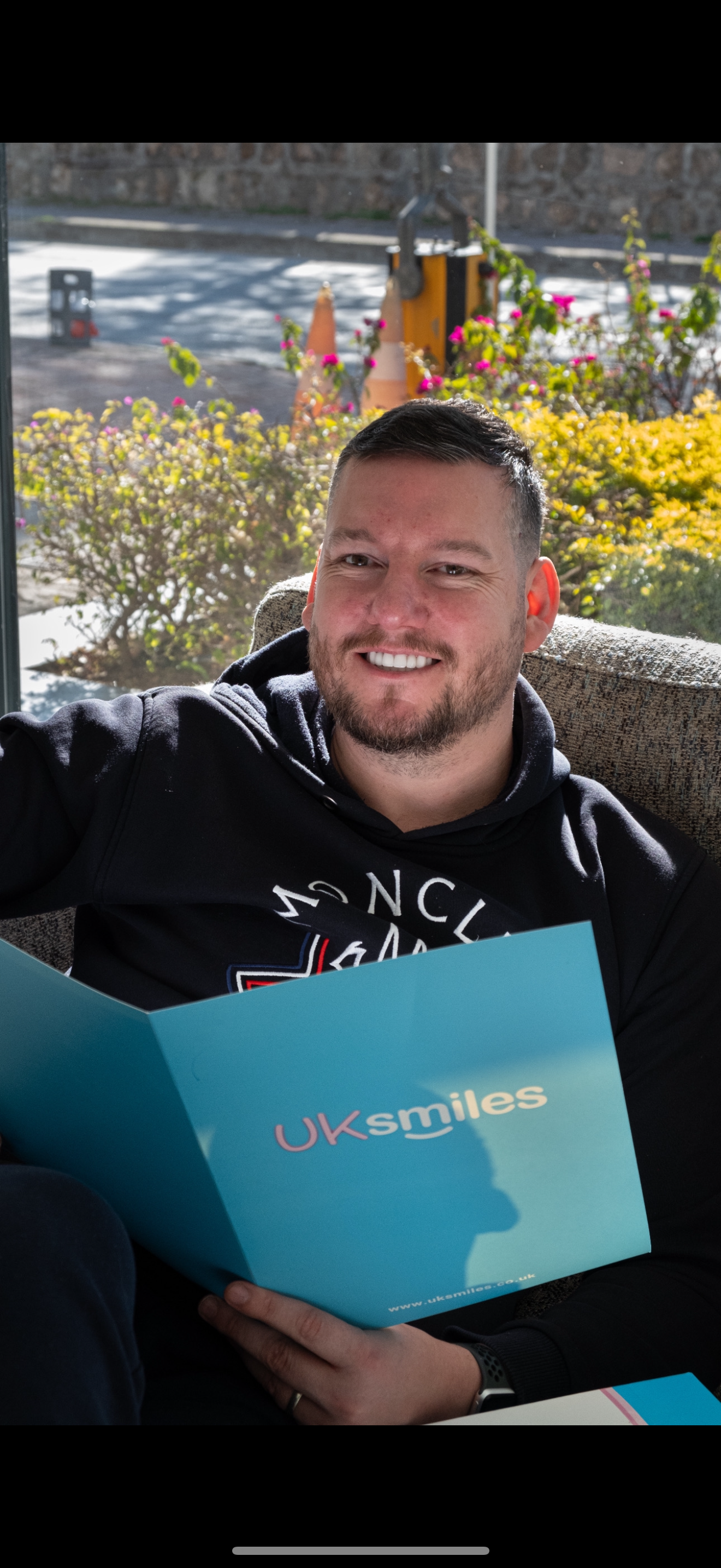 Man smiling, holding a blue brochure, sitting near a window, with a blurred outdoor view.