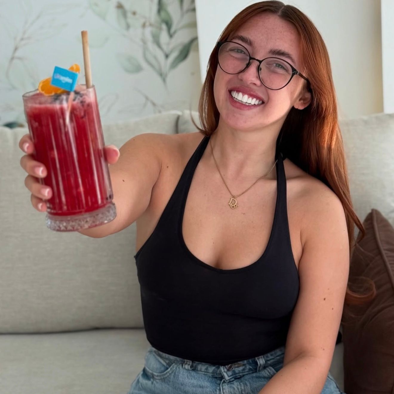 Woman smiles, holding a red drink with a straw. She wears a black top, glasses, and stands indoors.