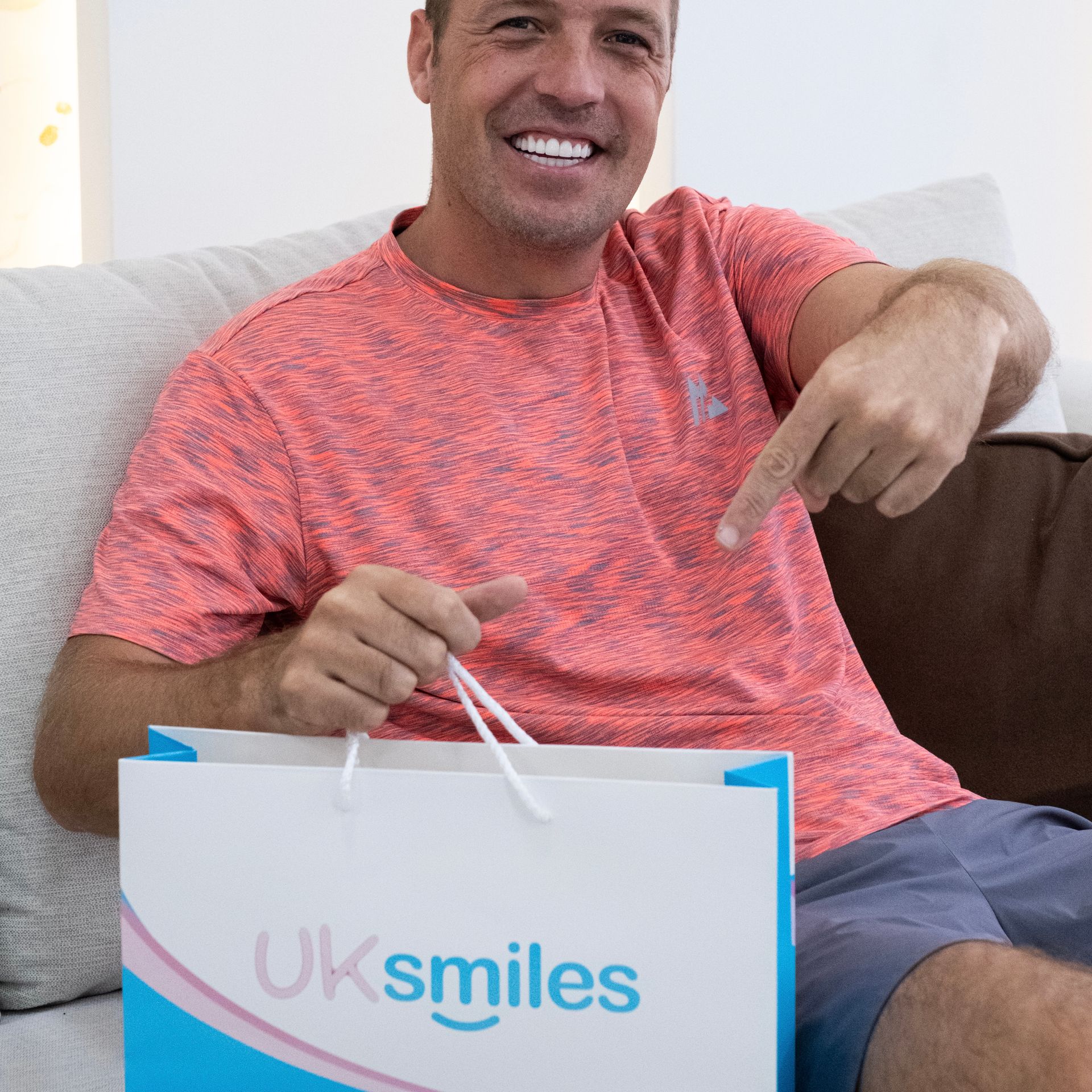 Man pointing at a UK Smiles shopping bag, smiling on a couch.
