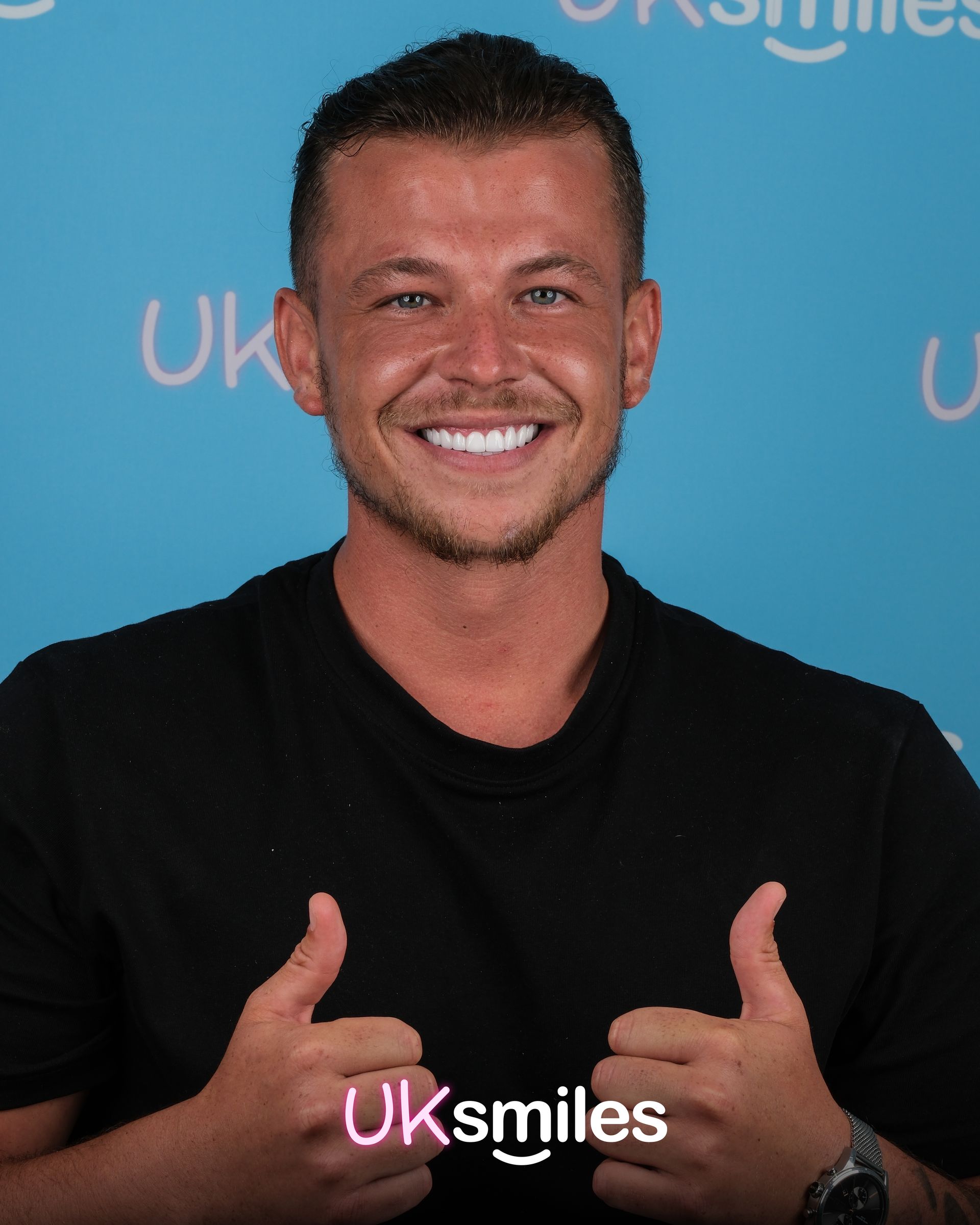 Man with white teeth, thumbs up, smiling. Wearing a black shirt, against a blue background. UKsmiles logo.