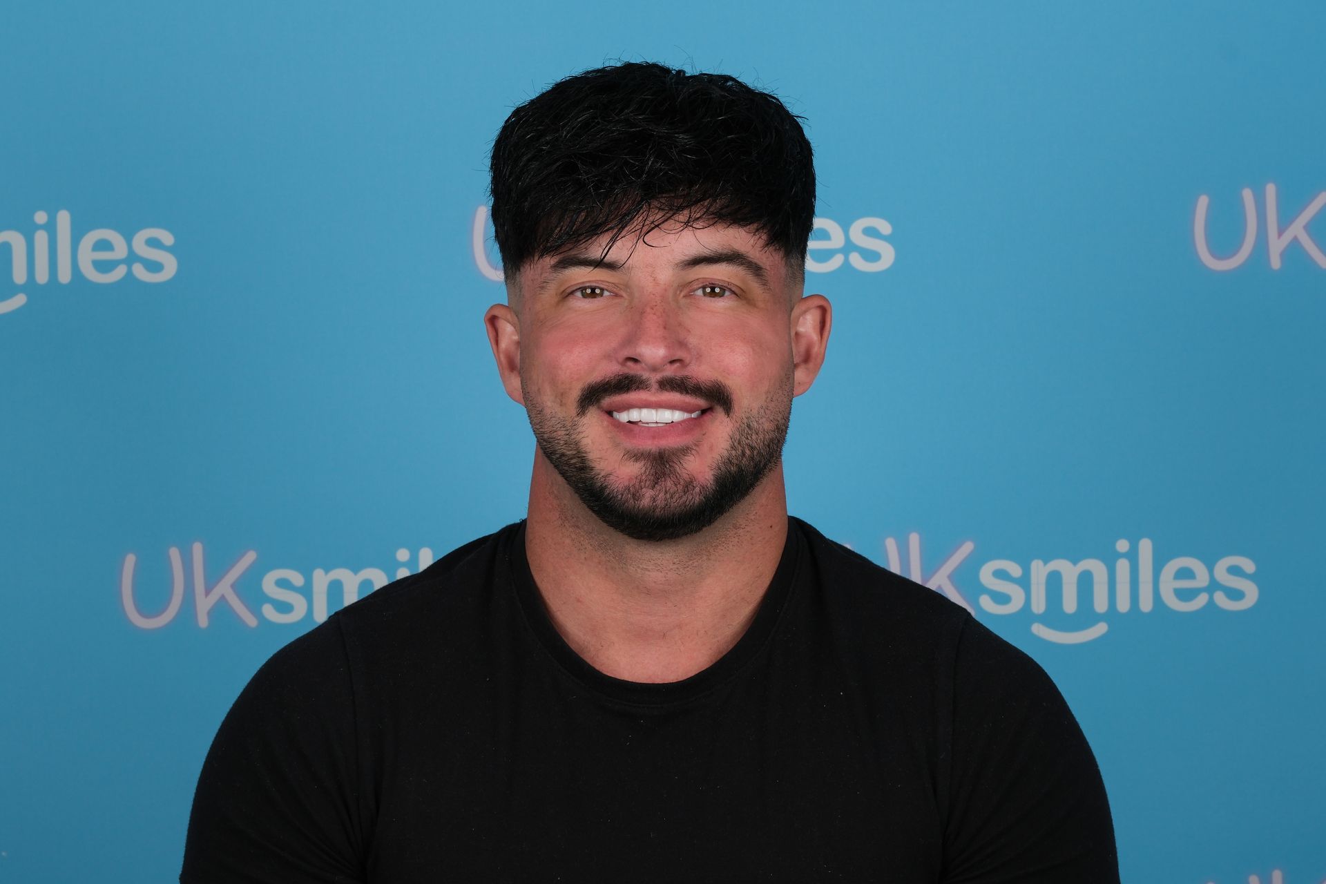 Man with dark hair and beard smiling at the camera in front of a blue backdrop.