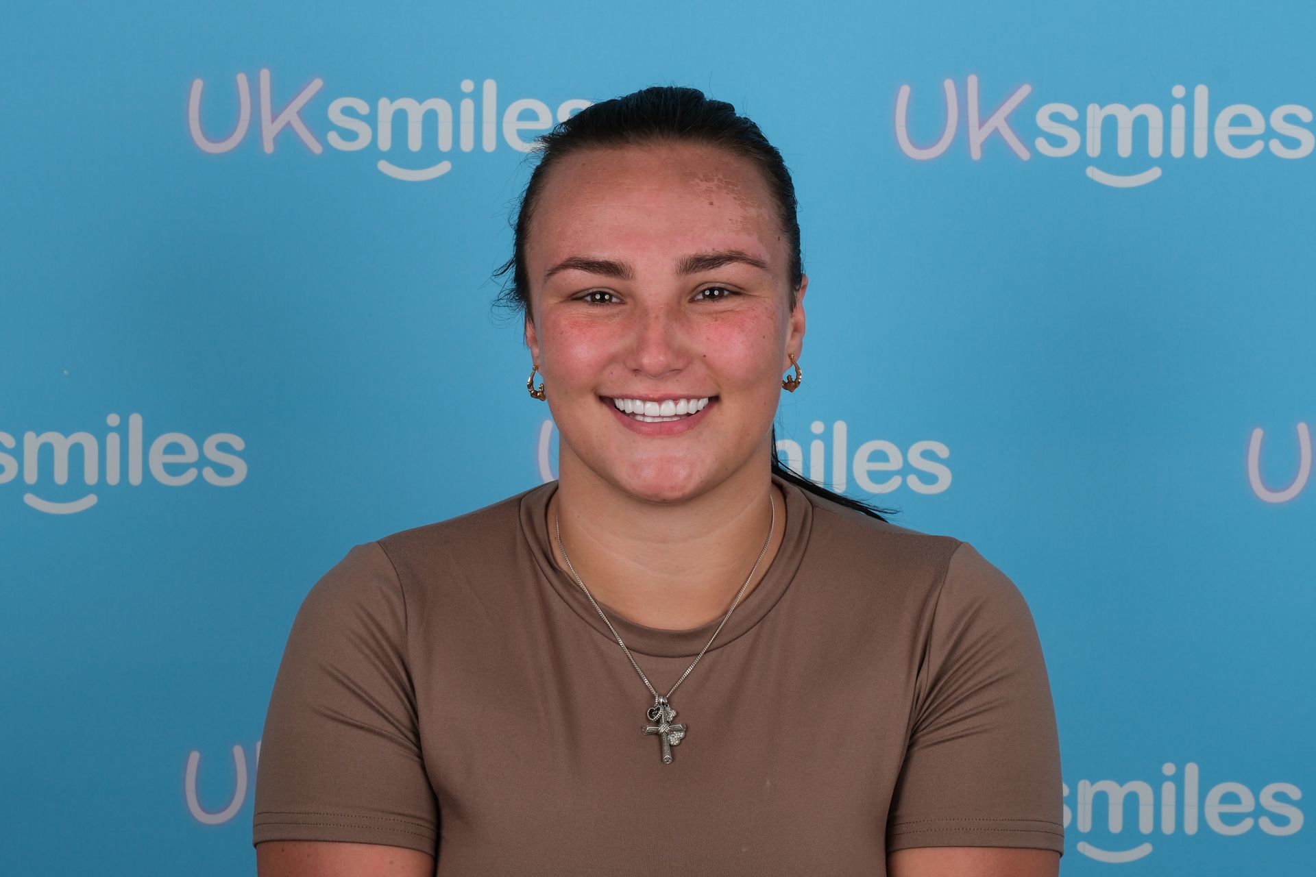 Woman with missing front tooth smiles at the camera, light brown shirt, blue background with
