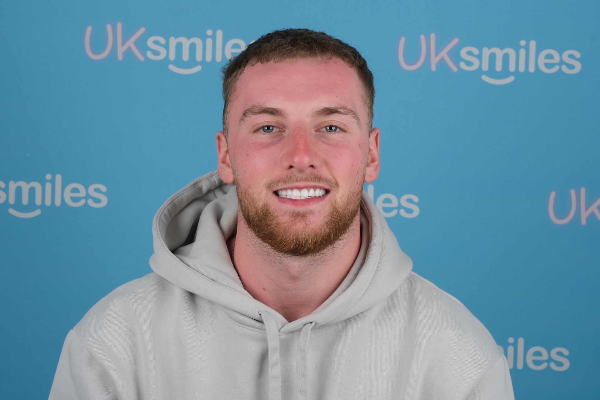 Man with light beard, wearing a gray hoodie, smiling with white teeth against a blue background.