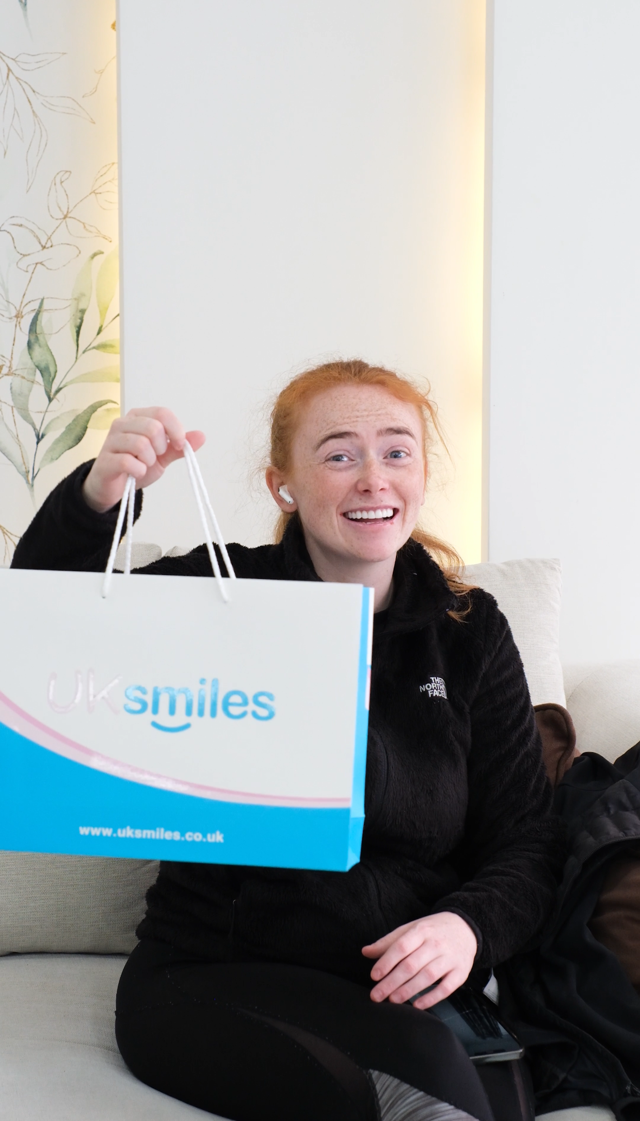 A woman with red hair smiles, holding a bag that says