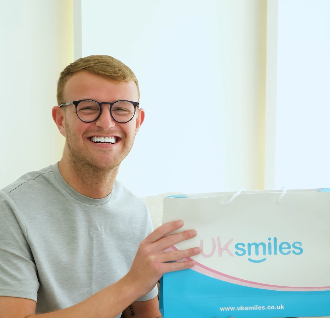 Man with glasses smiling, holding a UK smiles box. Interior setting.