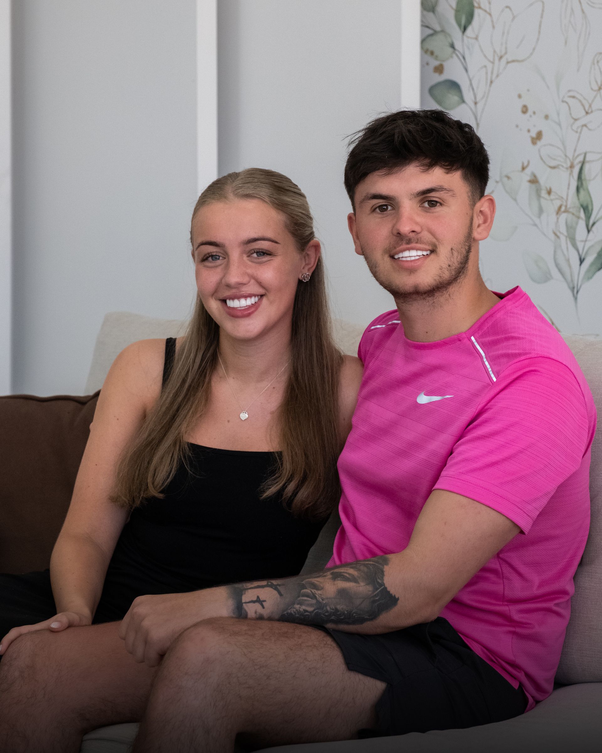 A young couple smiling on a couch. The woman wears a black top, and the man a pink shirt.