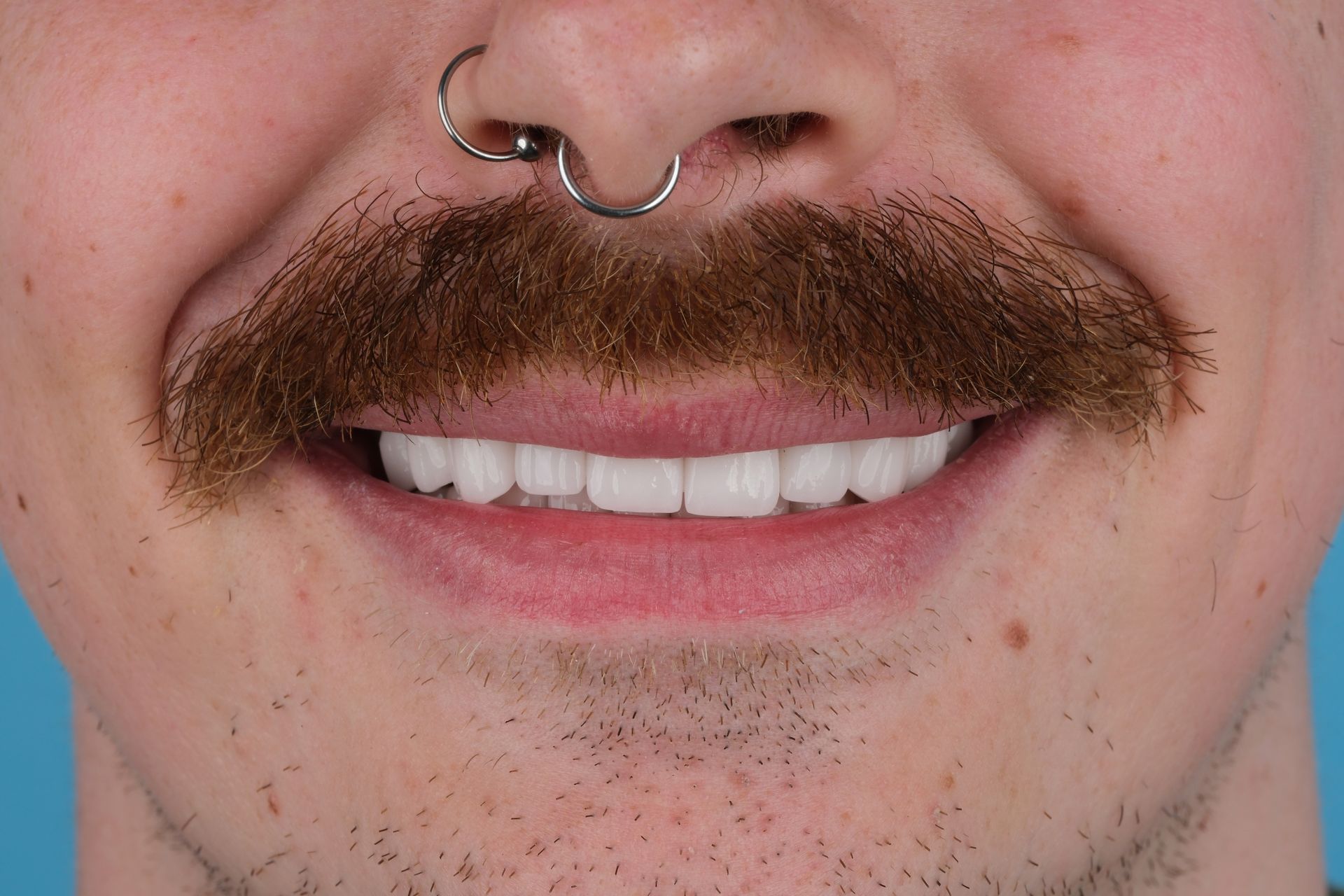 Close-up of a man smiling, showing white teeth, large brown mustache, and a nose ring.
