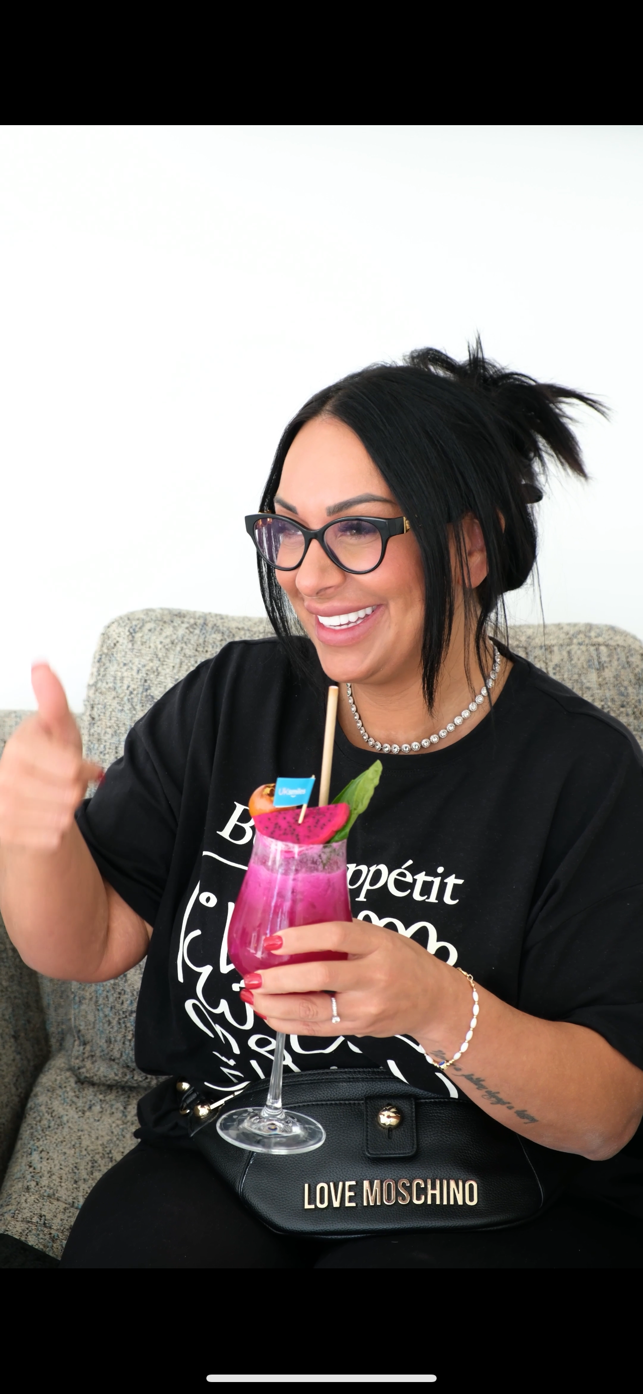Woman with glasses giving a thumbs-up while holding a pink cocktail; she smiles.