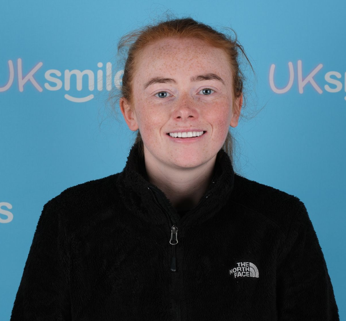 Woman with red hair, smiling, wearing a black jacket, in front of a blue backdrop with white text.