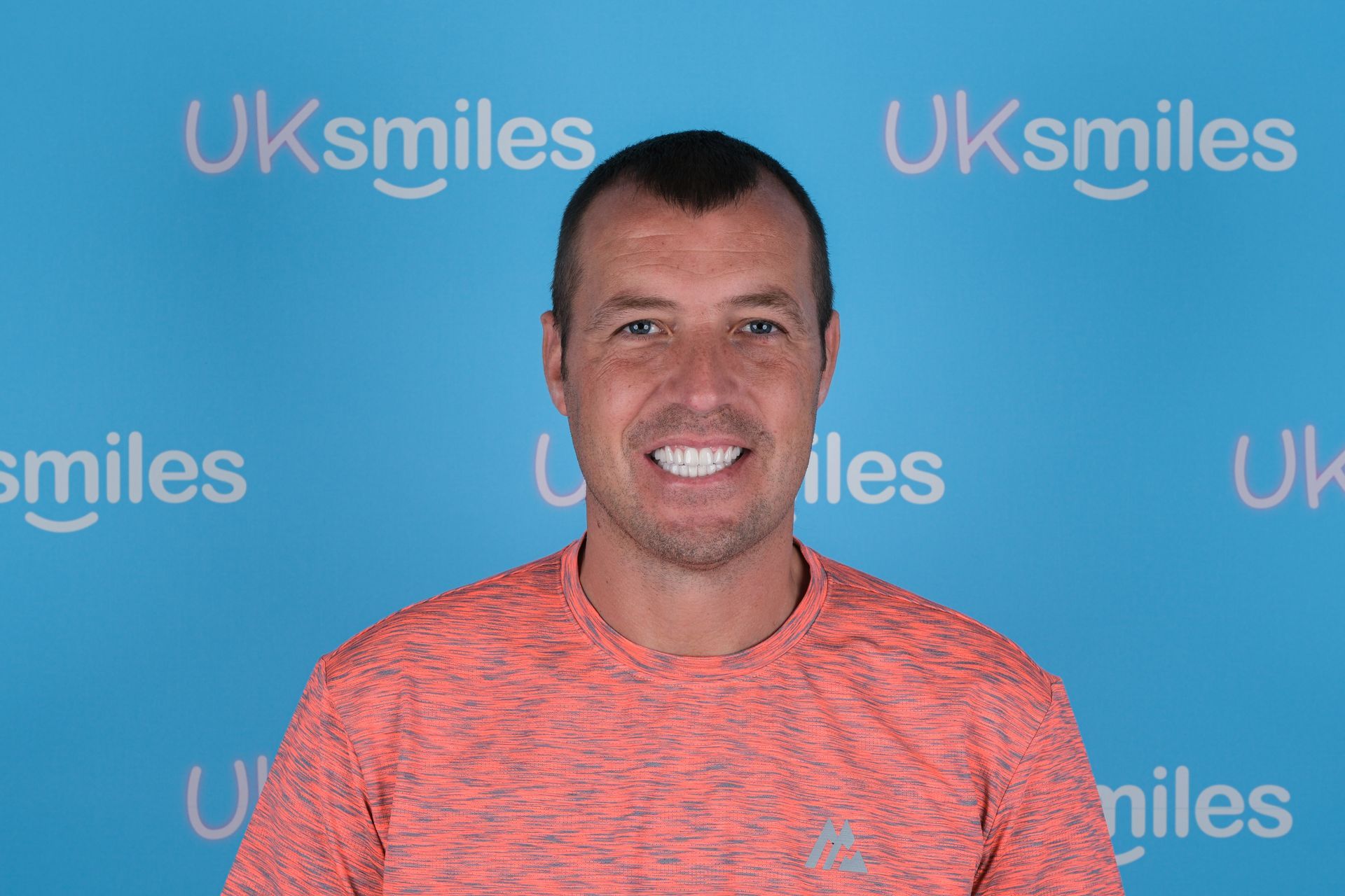 Man with a slight smile, wearing an orange shirt, in front of a blue background with