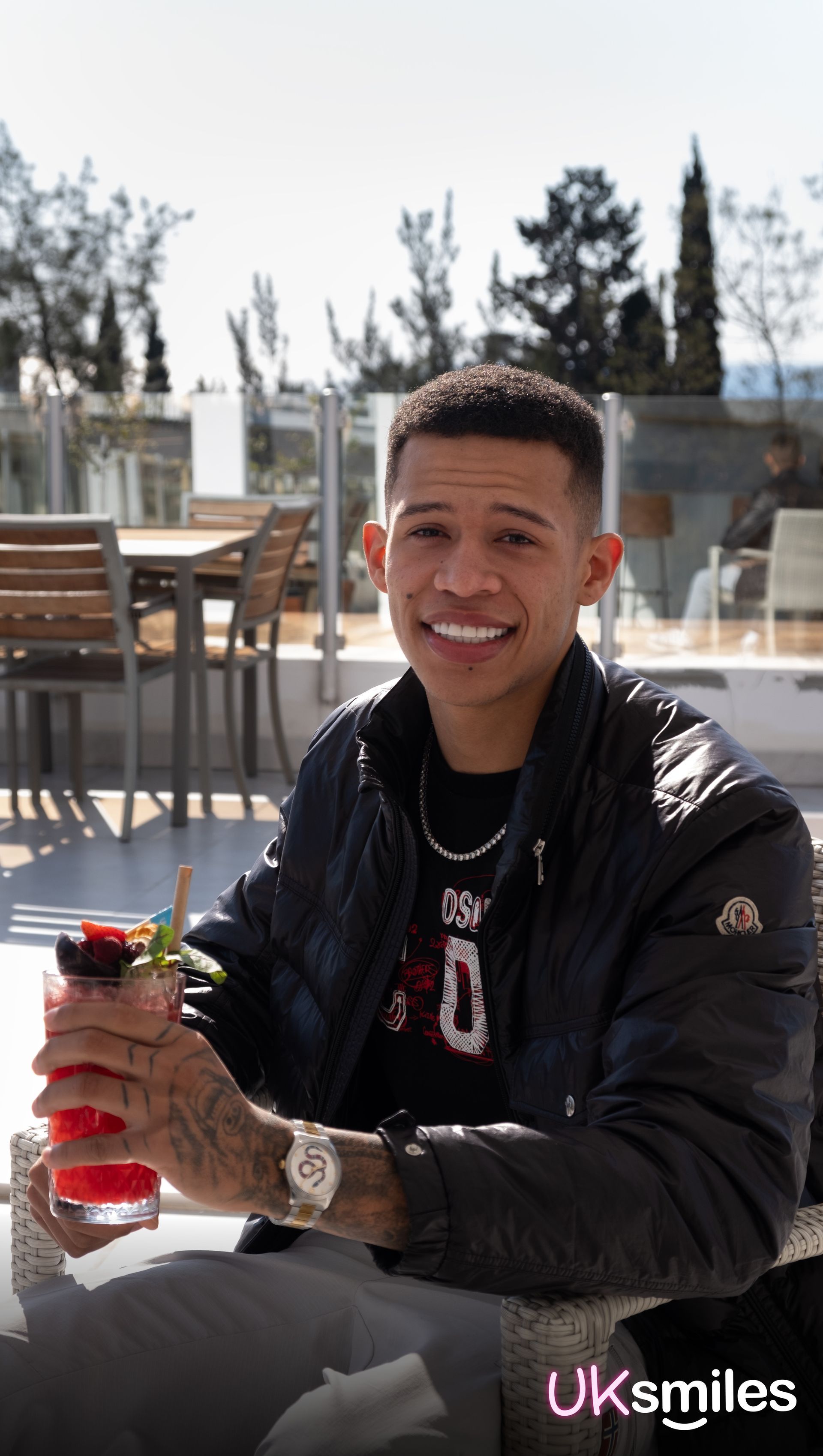 Man smiles, holding a drink, seated outdoors. Wearing black jacket, watch, necklace; visible tattoos. Bright teeth.